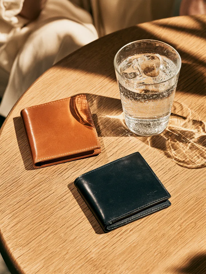 Two different minimalist wallets side by side on a light oak cafe table next to a glass of sparkling water